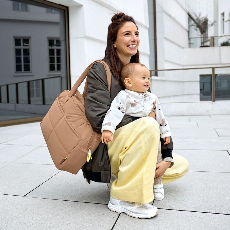 mutter mit baby auf schoß kniet auf boden große wickeltasche hellbraun über schulter